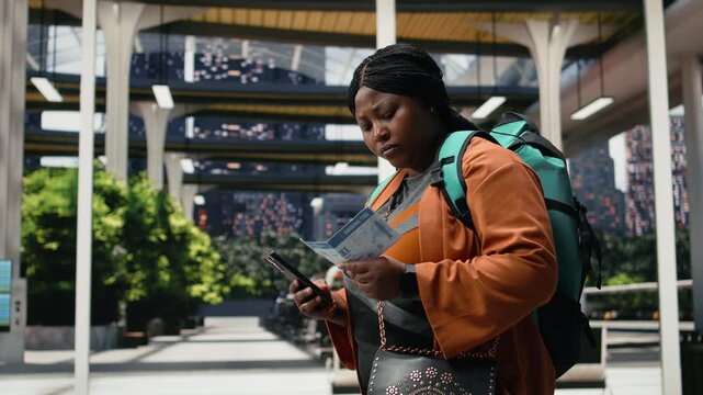 Smiling woman compares a printed bus ticket with information on a smartphone inside a terminal; the traveler checks her travel document and departure details on the coach platform.