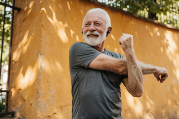 Obraz na płótnie Canvas Smiling elderly man with white hair and beard stretches arms outdoors. He wears earphones, listening to music while exercising in daylight near yellow wall and trees.