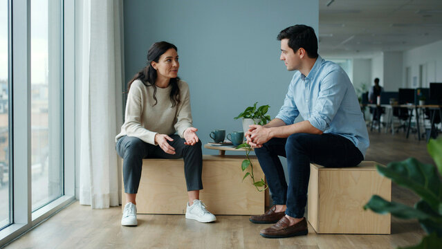 Two professional colleagues having a conversational meeting while sitting on wooden blocks in modern office with engaged conversation natural light streaming large window