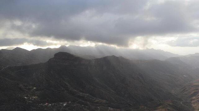 Aerial drone shot of the dramatic mountain landscape of Gran Canaria, Canary Islands. Sunrays break through clouds over Timagada village and rugged valleys as the drone pans left.
