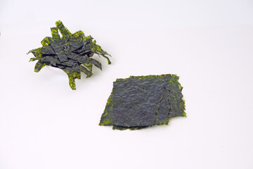 Fresh seaweed displayed on a clean white surface. One pile rests in a small bowl, slightly curled. Another lies flat, dark green with visible texture. Simple background highlights natural colors © Nurma