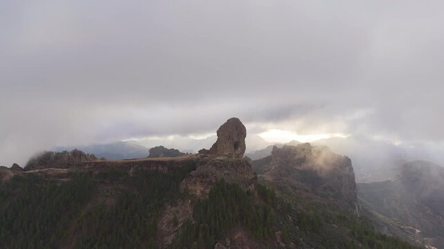 Aerial tracking shot panning left over the majestic Roque Nublo, an iconic volcanic pinnacle on Gran Canaria, Canary Islands, revealing rugged mountain scenery under an overcast sky.