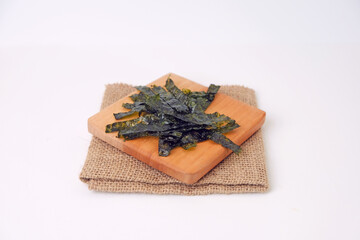 Dried seaweed rests on a wooden cutting board. Board sits atop a folded burlap napkin. White background offers clean, minimalist presentation. Seaweed leaves display natural texture and rich color © Nurma