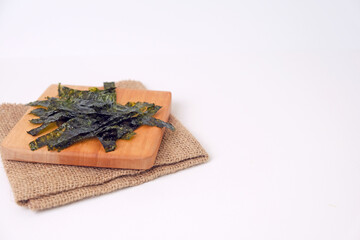 Dried seaweed rests on a wooden cutting board. Board sits atop a folded burlap napkin. White background offers clean, minimalist presentation. Seaweed leaves display natural texture and rich color © Nurma