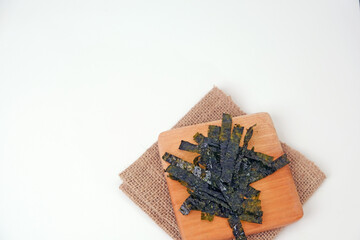 Dried seaweed rests on a wooden cutting board. Board sits atop a folded burlap napkin. White background offers clean, minimalist presentation. Seaweed leaves display natural texture and rich color © Nurma