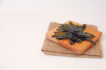Dried seaweed rests on a wooden cutting board. Board sits atop a folded burlap napkin. White background offers clean, minimalist presentation. Seaweed leaves display natural texture and rich color © Nurma