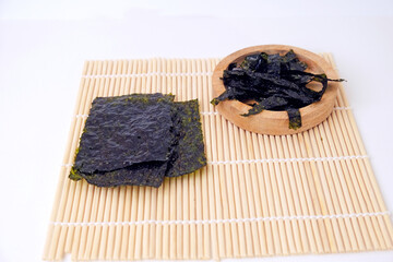 Kelp sheets rest on bamboo mat beside wooden bowl. Dried seaweed displays rich dark green hue. Simple background highlights natural textures and colors. Ideal for health, wellness © Nurma