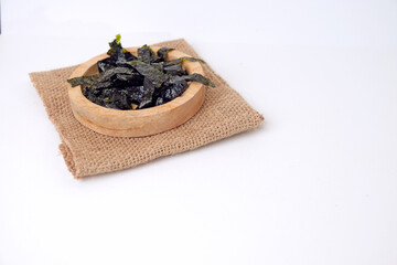 Dried seaweed rests in a wooden bowl. Burlap cloth provides rustic contrast beneath. Clean white background emphasizes simplicity and texture © Nurma