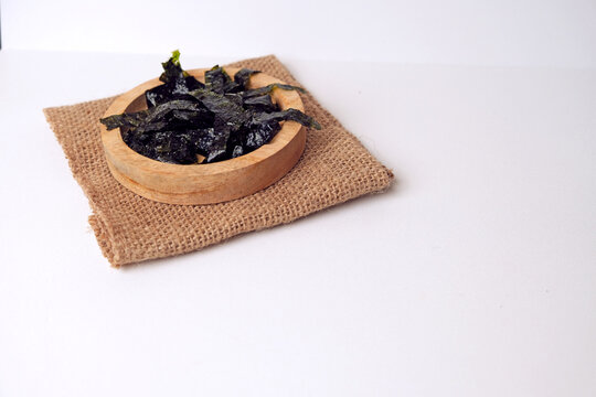 Dried seaweed rests in a wooden bowl. Burlap cloth provides rustic contrast beneath. Clean white background emphasizes simplicity and texture