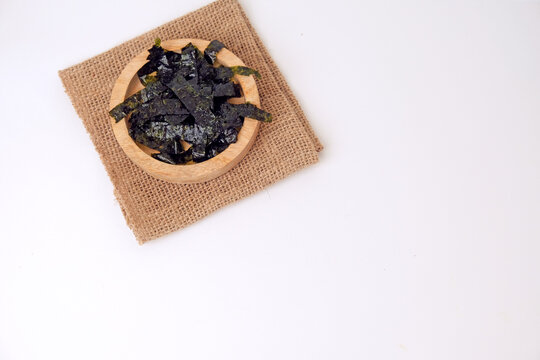 Dried seaweed rests in a wooden bowl. Burlap cloth provides rustic contrast beneath. Clean white background emphasizes simplicity and texture