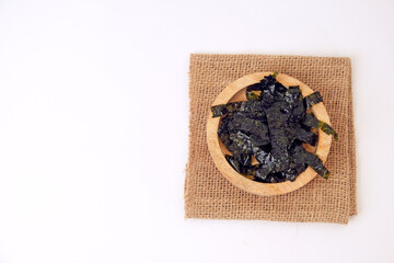 Dried seaweed rests in a wooden bowl. Burlap cloth provides rustic contrast beneath. Clean white background emphasizes simplicity and texture © Nurma