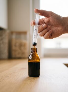 Woman dispensing dark liquid cough syrup with a plastic oral syringe into a medicine bottle on a kitchen counter