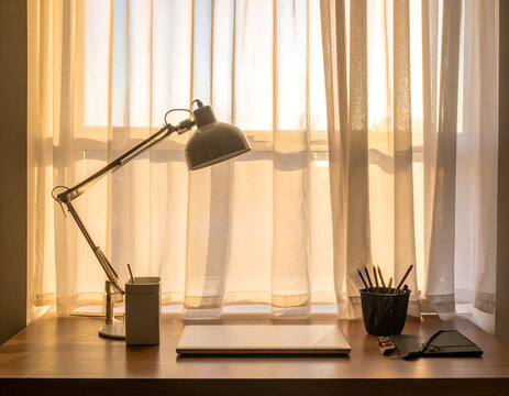 Desk lamp and closed laptop on a wooden table beside a sunlit window.