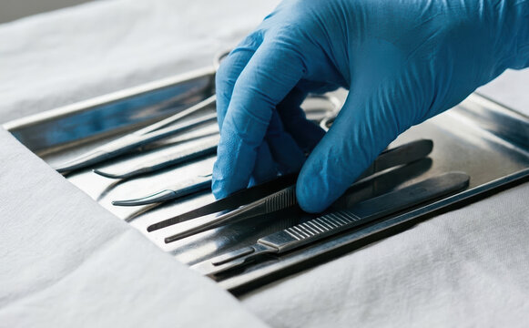 Surgeon in blue gloves picking up surgical tweezers from a metal tray in a sterile operating room