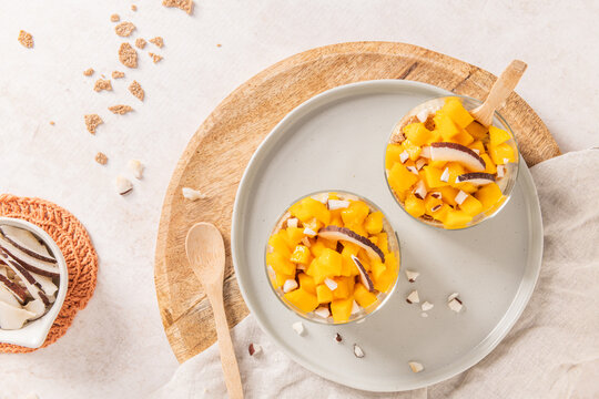 Overhead view of two glass jars filled with fresh mango chunks and coconut flakes on a round wooden board and grey plate surrounded by rustic linens and copy space for a bright healthy concept