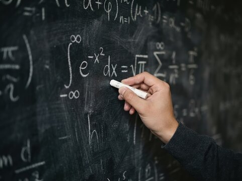 Hand of a young adult male writing mathematical formulas with chalk on a blackboard