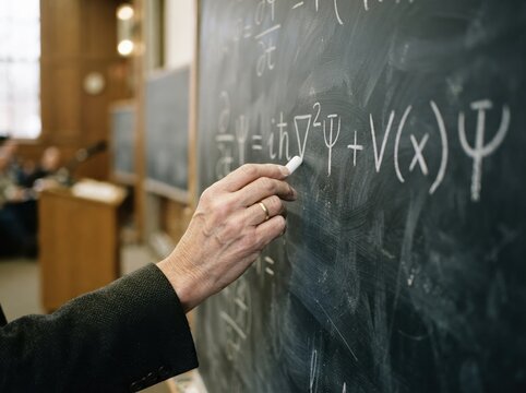 Elderly male professor writing quantum physics equations with chalk on a blackboard in a university lecture hall