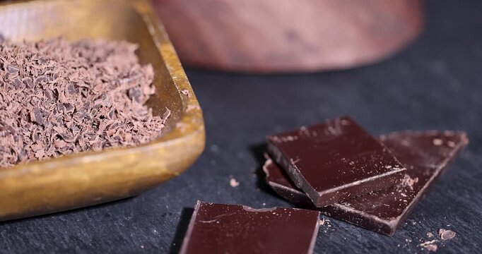 Brown grated chocolate in wooden bowl, dark chocolate pieces lying on black stone background table next to dessert ingredients