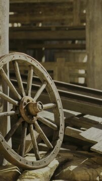 Weathered wooden wheel rests against a sturdy pole inside an old barn. Sunlight spills gently into the space, highlighting the timeless beauty of this rural scene.