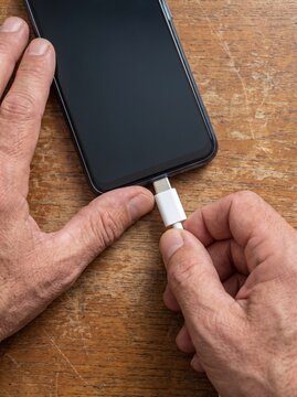Close up of hands plugging a white USB C cable into a modern smartphone charging port on a wooden table