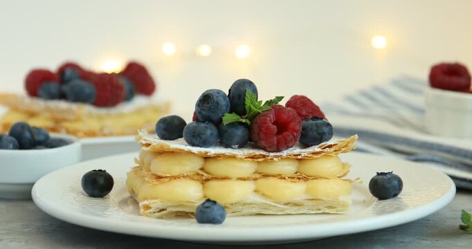 Woman putting blueberries onto mille-feuille with custard and berries on table, closeup. Puff pastry product