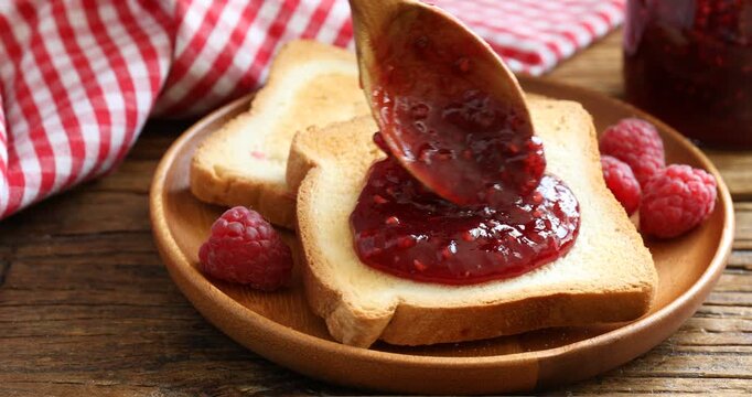 Spreading delicious raspberry jam onto toast at wooden table, closeup
