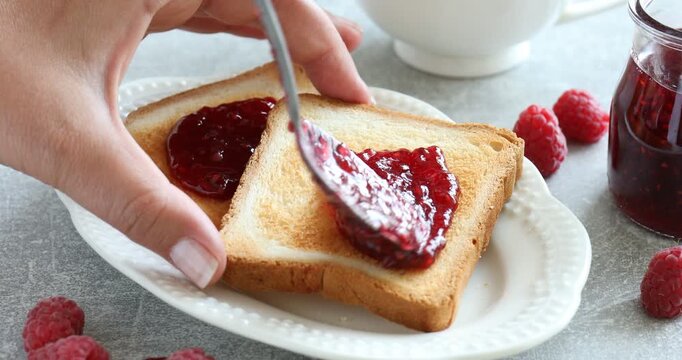 Woman spreading delicious raspberry jam onto toast at gray textured table, closeup
