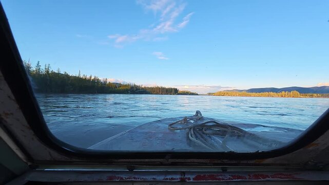 Front cockpit view of a river from a floatplane, surrounded by dense forest, distant mountains, under clear blue sky with scattered clouds.