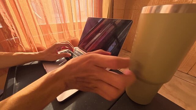 Gresik, Indonesia - August 4, 2026: Side angle view of a person typing on an Apple MacBook laptop near a window with beautiful morning sunlight streaming through curtains, enjoying a drink at home