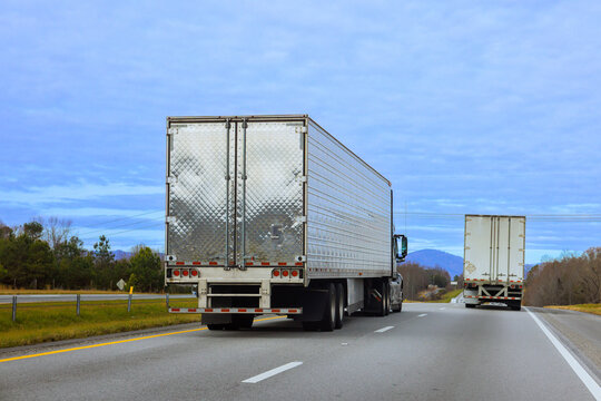 Two large trucks are moving along highway surrounded by trees mountains in distance.