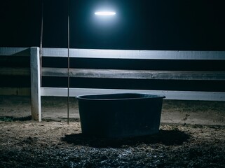 Fototapeta premium A black rubber feed tub is positioned next to a wooden fence, illuminated by an overhead light in the darkness of night