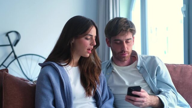 Millennial couple sitting together on a sofa in their living room, intently looking at a mobile phone screen and talking, showing different emotions from interest to disagreement and disappointment