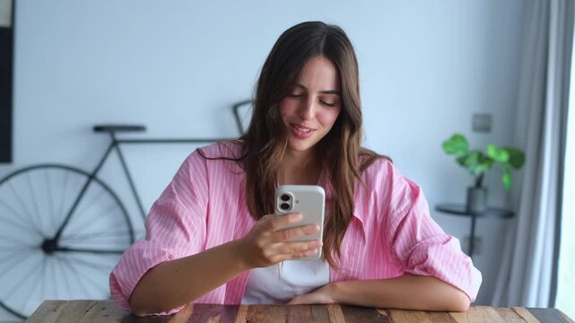 Beautiful smiling woman using her mobile phone while sitting at a wooden table in her modern apartment, enjoying online communication, browsing social media, and looking thoughtfully out the window