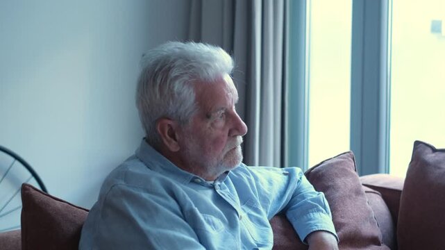 Solitary senior man with white hair sitting on a couch at home, looking out the window with a pensive and melancholic expression, feeling lonely and isolated while thinking about life