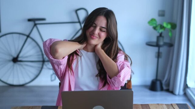 Overworked young woman feeling tired and suffering from neck pain due to a sedentary lifestyle, incorrect posture, and long hours working on her laptop in a modern home office interior