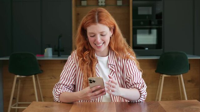 Beautiful young woman with long red hair smiling while using her smartphone, scrolling through social media, and typing messages while sitting at a table in her modern kitchen