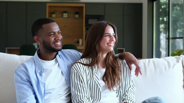 Young happy interracial couple sitting on a sofa, looking away and dreaming of a shared future. Smiling african american husband hugging his beautiful caucasian wife, relaxing at home