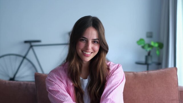 Portrait of a charming young woman with long brown hair, sitting on a couch in her modern living room and smiling sweetly at the camera, conveying happiness and contentment at home