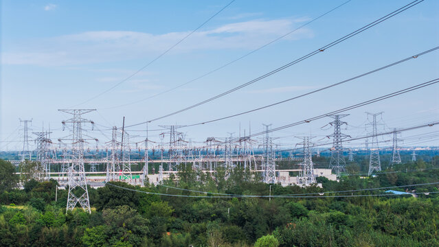 An outdoor high voltage power substation with multiple transmission towers and power lines, surrounded by green vegetation under a clear sky.