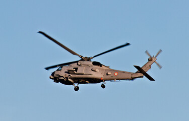 Fototapeta premium A naval military helicopter flies through a clear blue sky with landing gear lowered during an apparent coastal aviation operation.