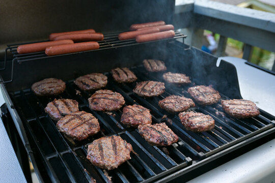 Hamburger patties and hot dogs cooking on a smoky outdoor barbecue grill during a backyard cookout.