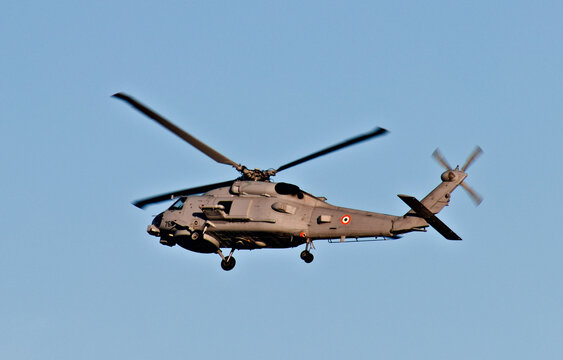 A naval military helicopter flies through a clear blue sky with landing gear lowered during an apparent coastal aviation operation.