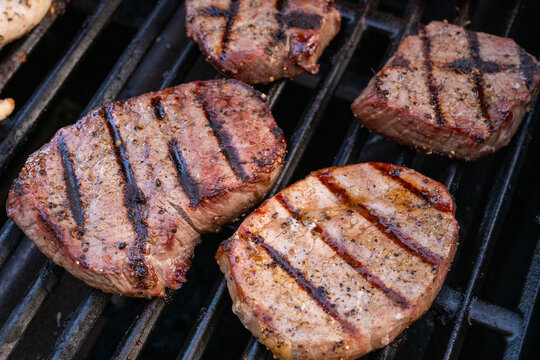 Close-up photo of seasoned steaks cooking on a barbecue grill with dark char marks and a smoky outdoor cooking look.