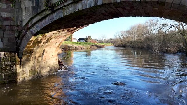 Smooth low altitude drone flight beneath a historic stone arch bridge over a peaceful river in rural England. Scenic countryside landscape with calm water and traditional architecture captured in cine