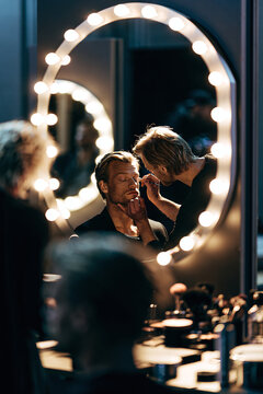Professional makeup artist applying cosmetics to male actor in illuminated vanity mirror backstage