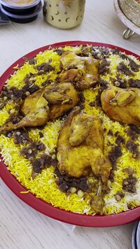 High angle close-up of a traditional Kuwaiti chicken machboos platter on a wooden dining table. The large red tray features yellow rice, roasted chicken, and hashwu onions in a bright indoor setting.