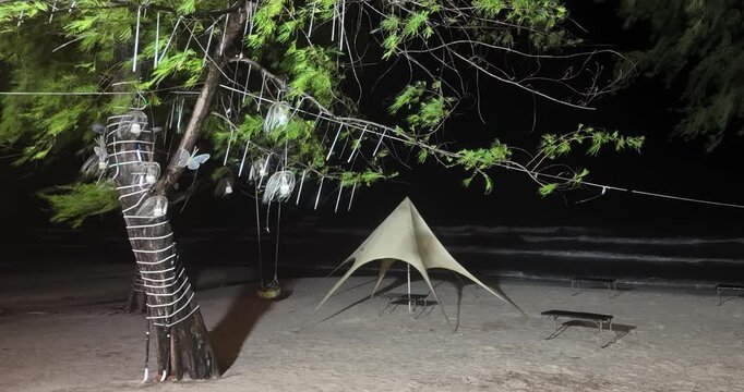 Beach scene with waves and wind under rain, featuring a tree decorated with lights and butterfly ornaments, a star-shaped camping tent, and tables set on the sand against the dark ocean backdrop