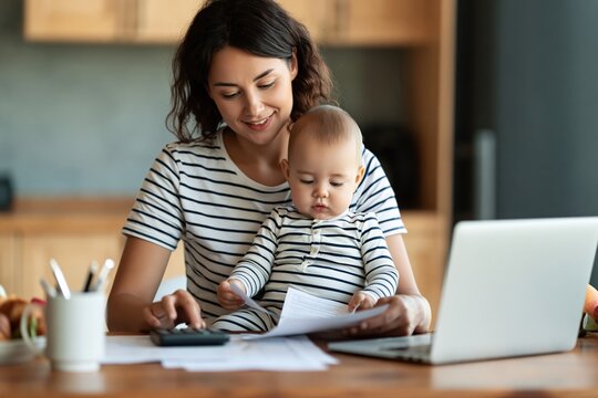 Mother multitasking with baby while working on laptop and calculator at home