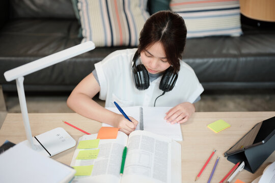 Asian teenage girl in headphones concentrates on learning at home, reading textbooks, note writing, self-tutorial exams, preparation for back to school, Homeschooling, university education lifestyle.