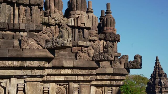 Carved temple wall leads toward a distant Prambanan spire under brilliant tropical sunlight and cloudless blue sky.